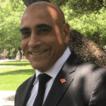 Building Safer Communities Together Active Shooter Prevention Project “Portrait of a professional man smiling in a dark suit, white shirt, and black tie, wearing a small lapel pin, with a neutral light background. Building Safer Communities Together Active Shooter Prevention Project